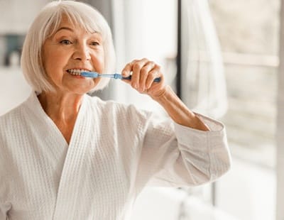 An older woman wearing a white bathrobe and brushing her teeth and dental implants